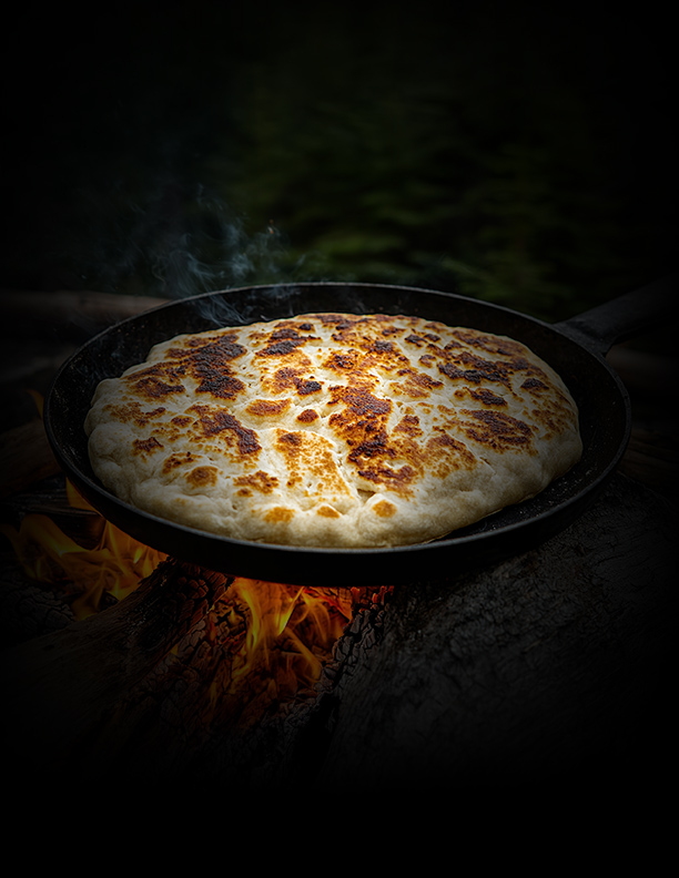 A round, golden-brown flatbread cooking in a black skillet over an open flame, with smoke rising and a dark, blurred background.